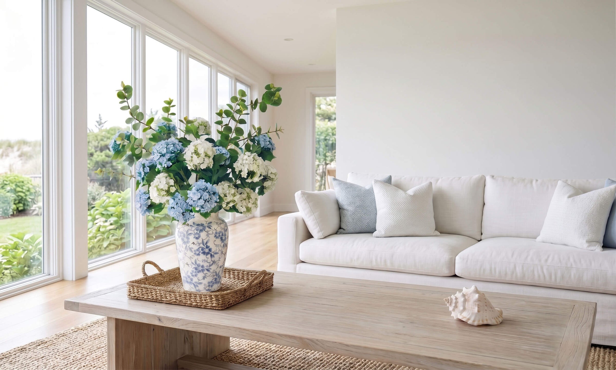 Artificial hydrangea arrangement in a blue and white porcelain vase displayed on a coffee table in a bright Hamptons coastal living room.